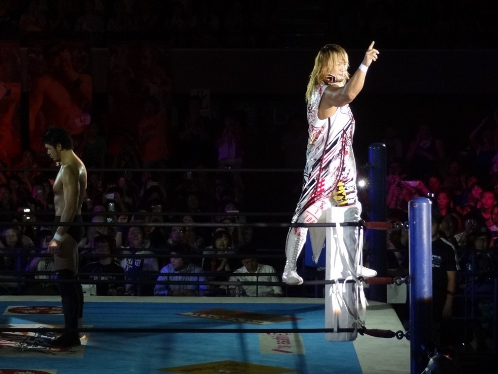 Hiroshi Tanahashi standing on the turnbuckle in the spotlight while Katsuyori Shibata stands unflinching in the shadows. 